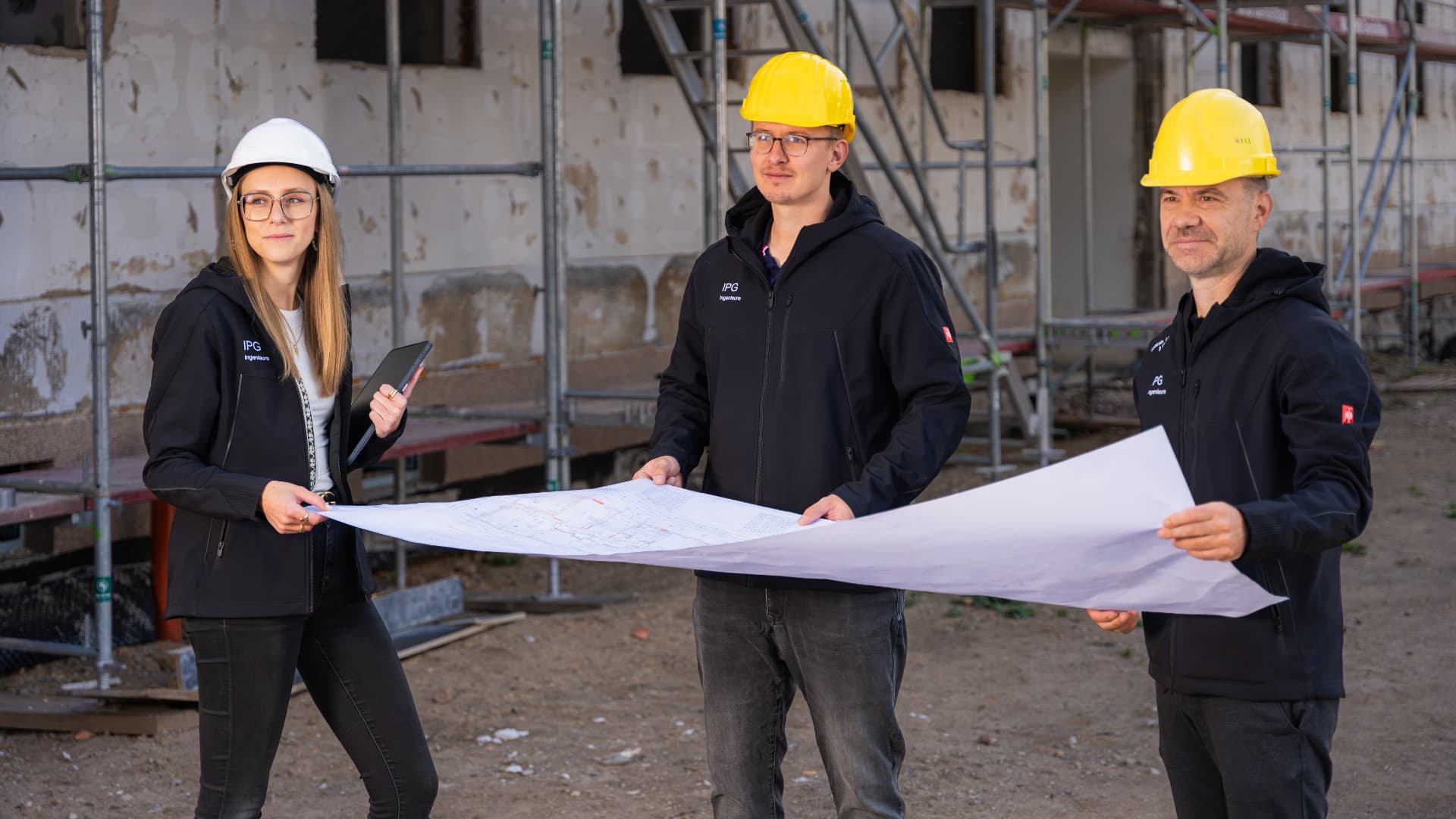 Drei Kolleginnen und Kollegen mit Schutzhelmen und Bauplan auf der Baustelle