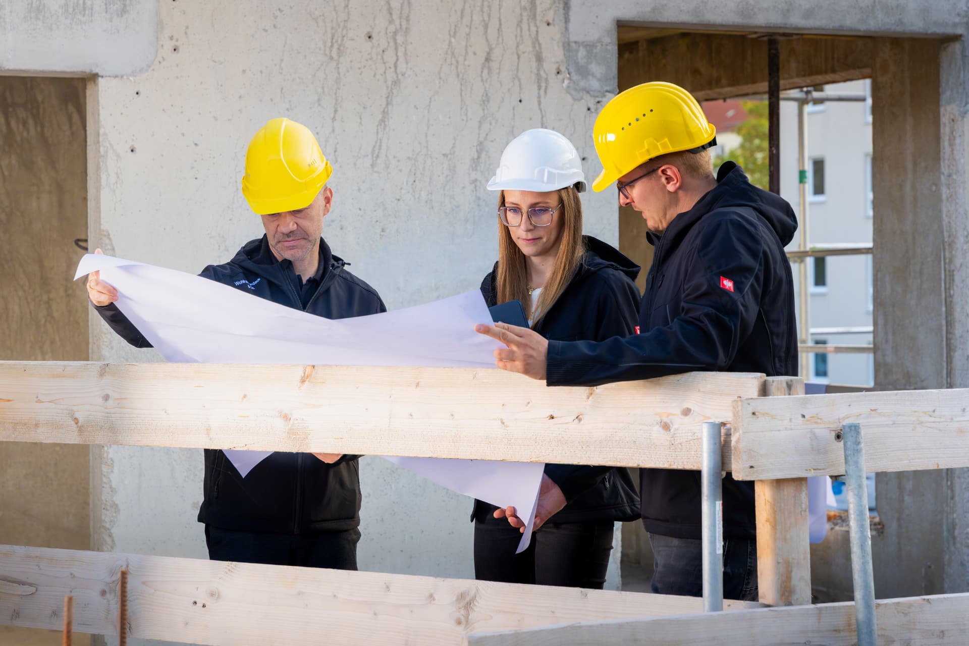 Drei Kolleginnen und Kollegen mit Schutzhelmen besprechen Pläne auf der Baustelle